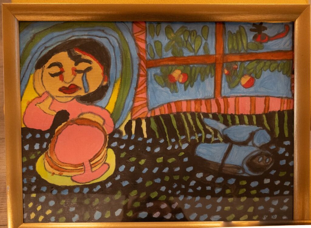 A black-haired woman with a sad face, a hand on her cheek and a blue tear streaming from her eye, sits at a table. A layered cake is placed on a plate on a brown tablecloth dotted with azure, yellow and greenish dots. Around the woman’s head are bluish stripes like a halo. Behind it, a window looks out onto branches with orange fruit.
