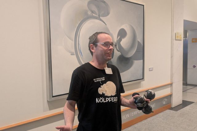 A man with short gray hair and glasses stands in front of a piece of art on the wall. He's wearing a black t-shirt and is holding four round objects in his left hand.