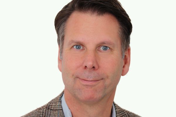 A man with short brown hair wears a light blue shirt and a checked jacket