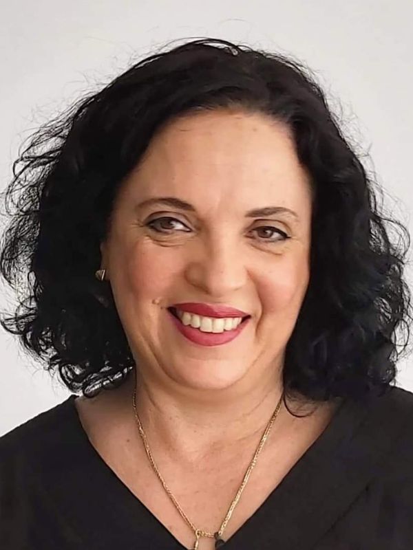 A smiling woman with brown curly hair and brown eyes wears a gold necklace and a black shirt.