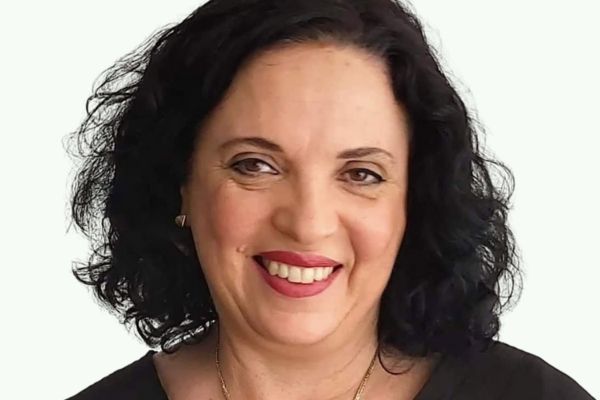 A smiling woman with brown curly hair and brown eyes wears a gold necklace and a black shirt.
