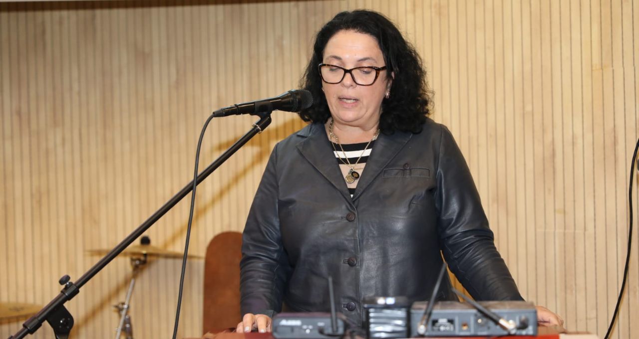 A woman with brown curly hair, brown glasses, and a dark leather jacket stands at the microphone behind a desk.