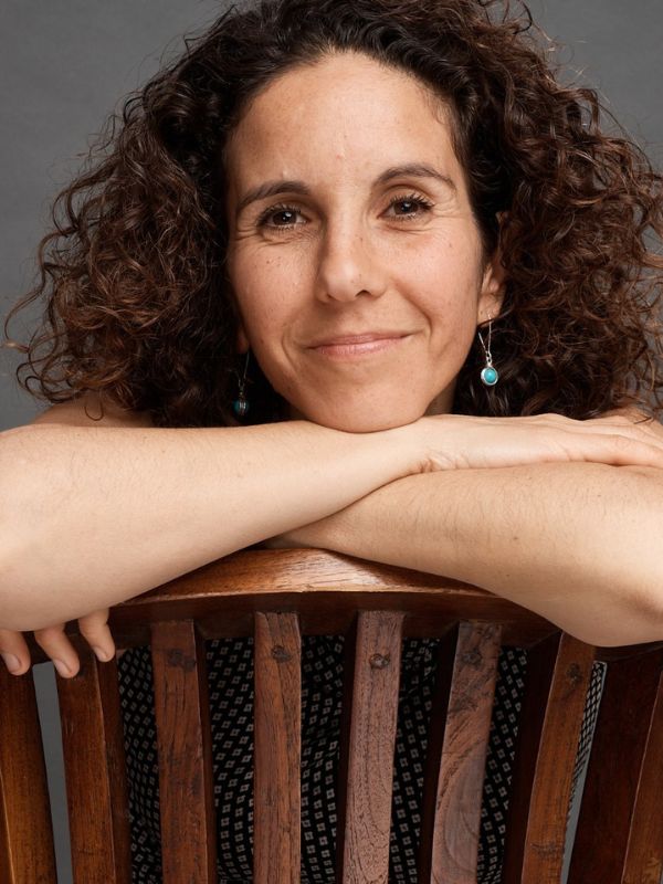 Vered Sabag A smiling woman with brown curly hair sits on a chair with her arms resting on the backrest.