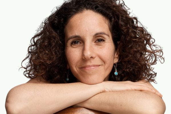 A smiling woman with brown curly hair sits on a chair with her arms resting on the backrest.