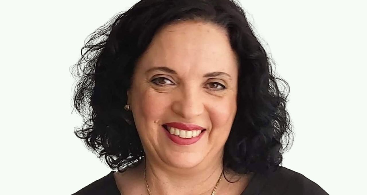 A smiling woman with brown curly hair and brown eyes wears a gold necklace and a black shirt.