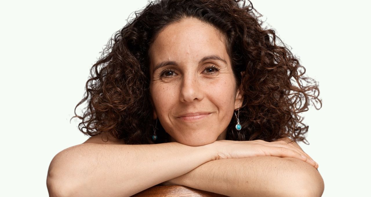 A smiling woman with brown curly hair sits on a chair with her arms resting on the backrest.